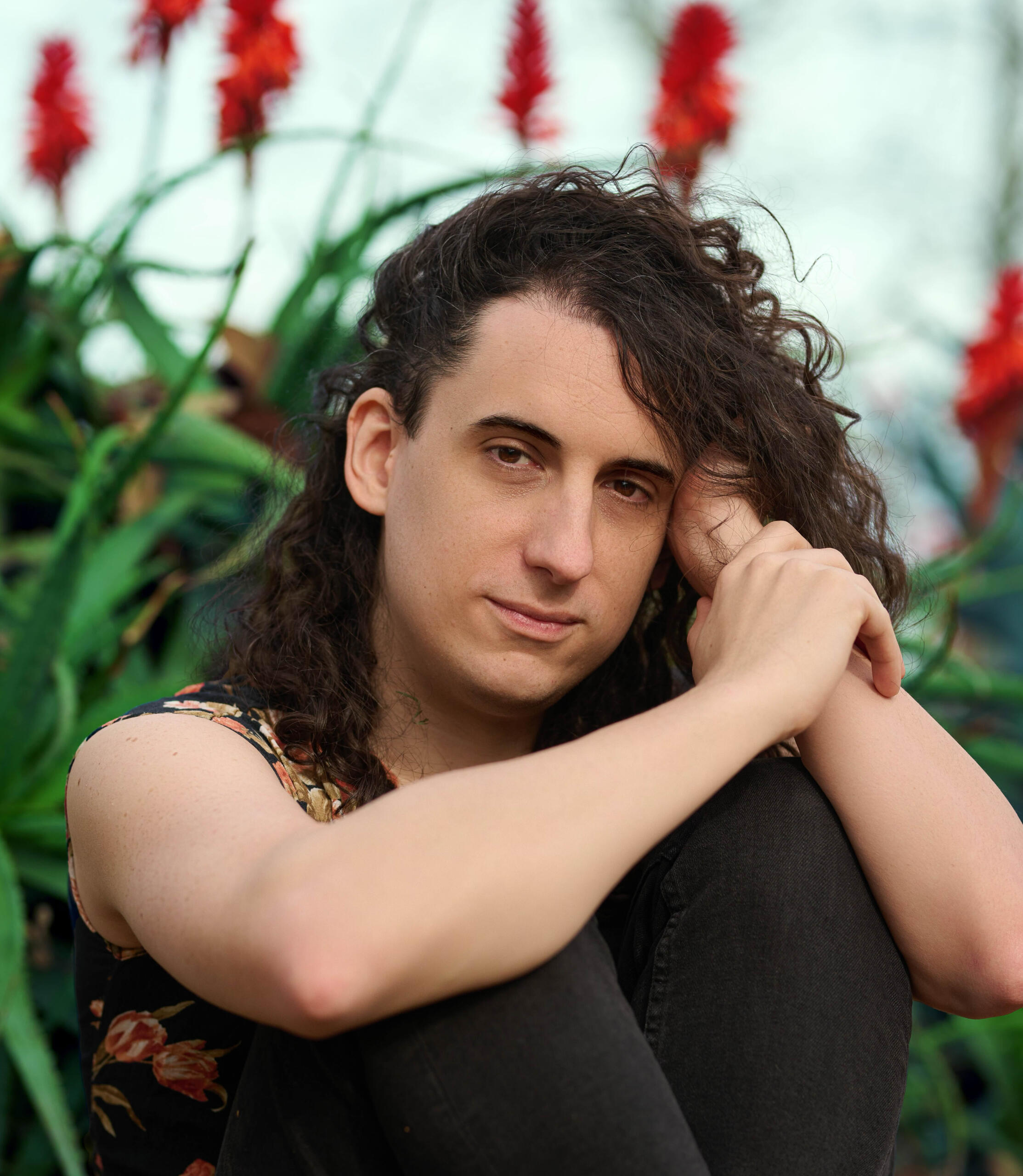 Melian Radu White femme with long curly dark hair, wearing a sleeveless floral top and black pants, sitting outdoors with knees up and arms resting on them, surrounded by green foliage and red flowers.